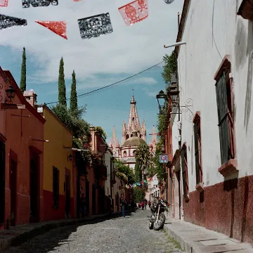 Cobblestone street lined with colorful buildings leading to a church with a tall spire, decorated with papel picado overhead.