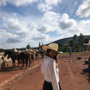 Woman wearing a white shirt and straw hat looking back, standing on a dirt path with several horses and a person in the background under a cloudy sky.