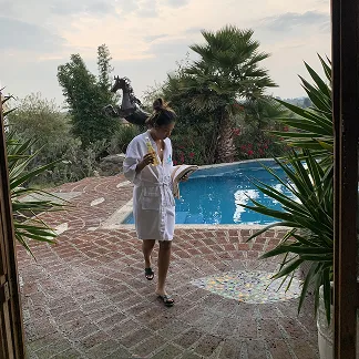 Person in a white bathrobe walking by a curved pool with palm trees and greenery in the background.