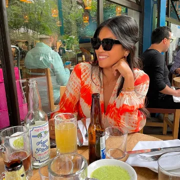 Woman with long dark hair wearing sunglasses and a red and white patterned blouse sitting at a restaurant table with drinks and food.