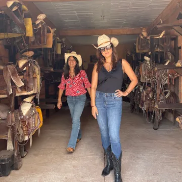 Two women wearing cowboy hats standing in a barn aisle flanked by hanging saddles on both sides.
