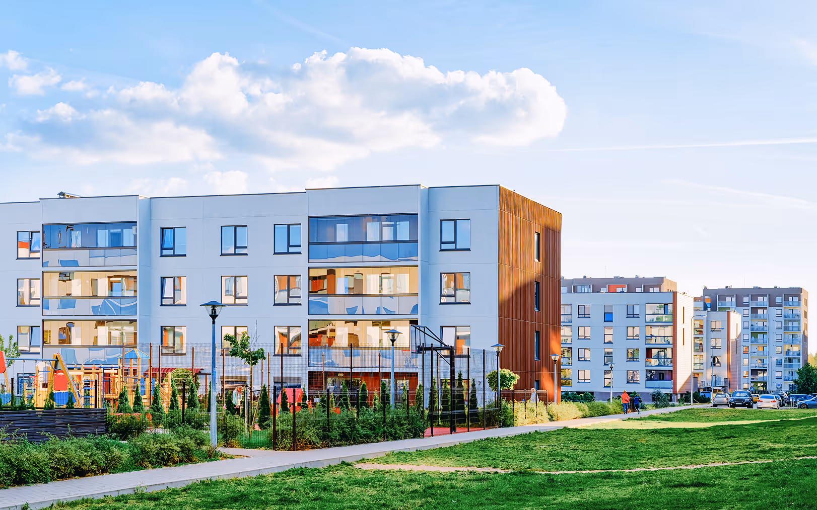 Moderne Wohngebäude mit Glasfenstern und Holzverkleidung neben einem Kinderspielplatz und einem Weg in einer grünen Anlage.
