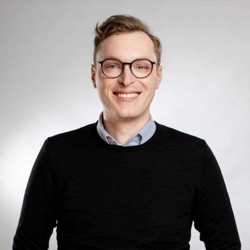 Porträt eines lächelnden Mannes mit Brille, blauem Hemd und schwarzem Pullover vor hellem Hintergrund.