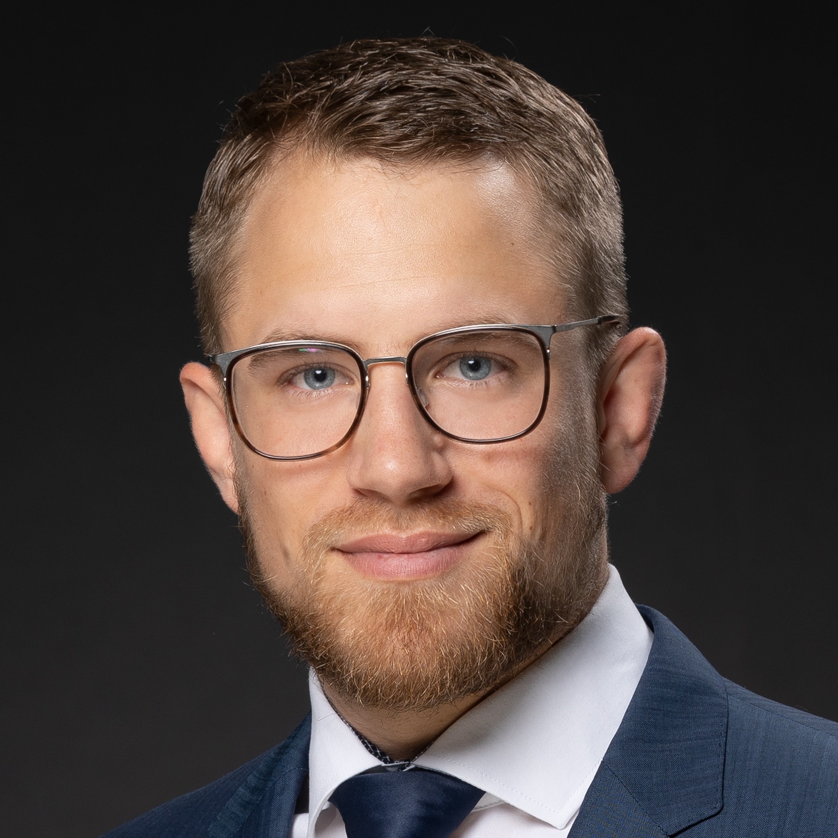 Porträt eines lächelnden Mannes mit kurzem braunem Haar, Bart und Brille, bekleidet mit Anzug, weißem Hemd und dunkelblauer Krawatte vor schwarzem Hintergrund.