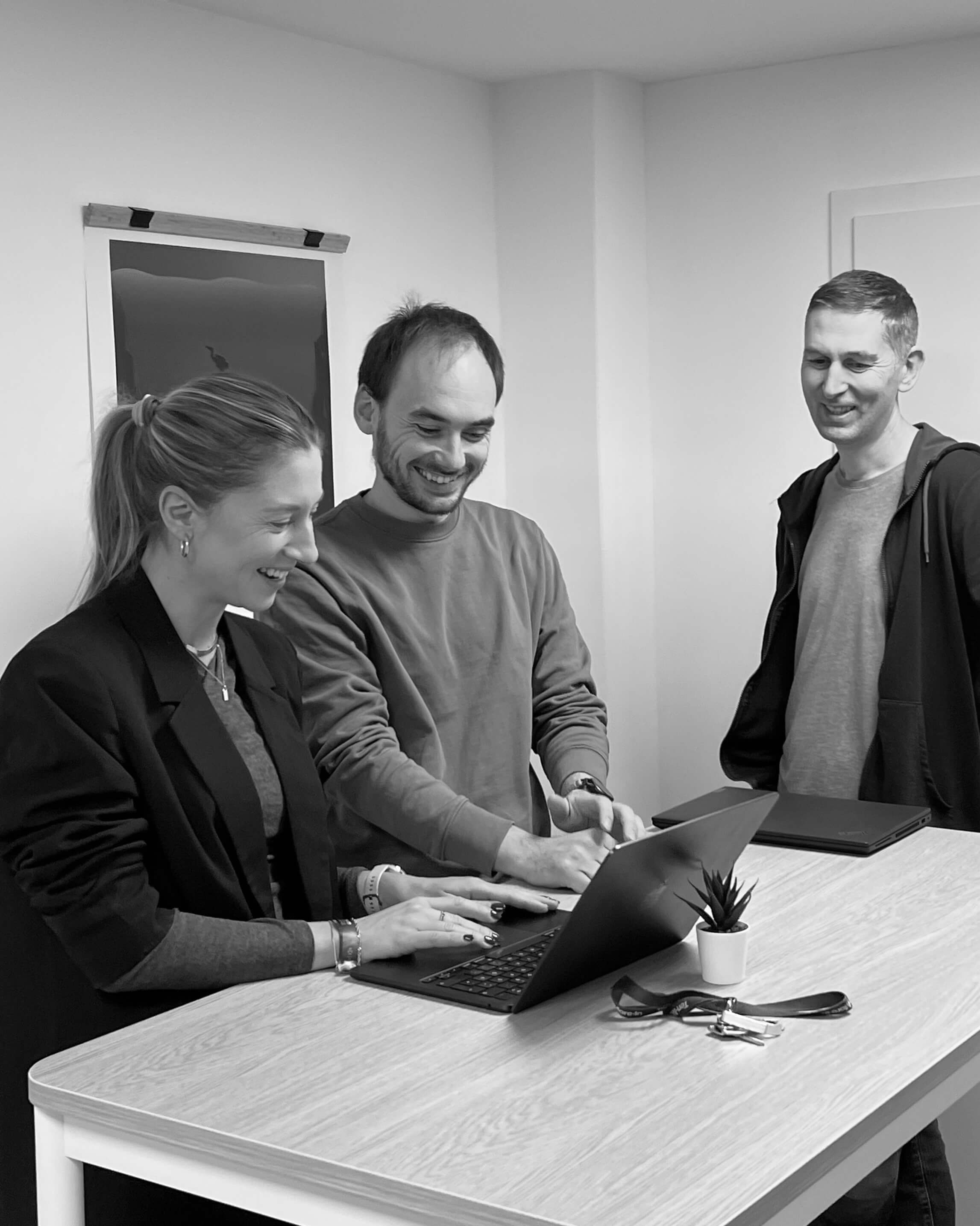 Drei lächelnde Personen stehen um einen Tisch, zwei bedienen einen Laptop, auf dem Tisch steht eine kleine Pflanze und ein Schlüsselbund.