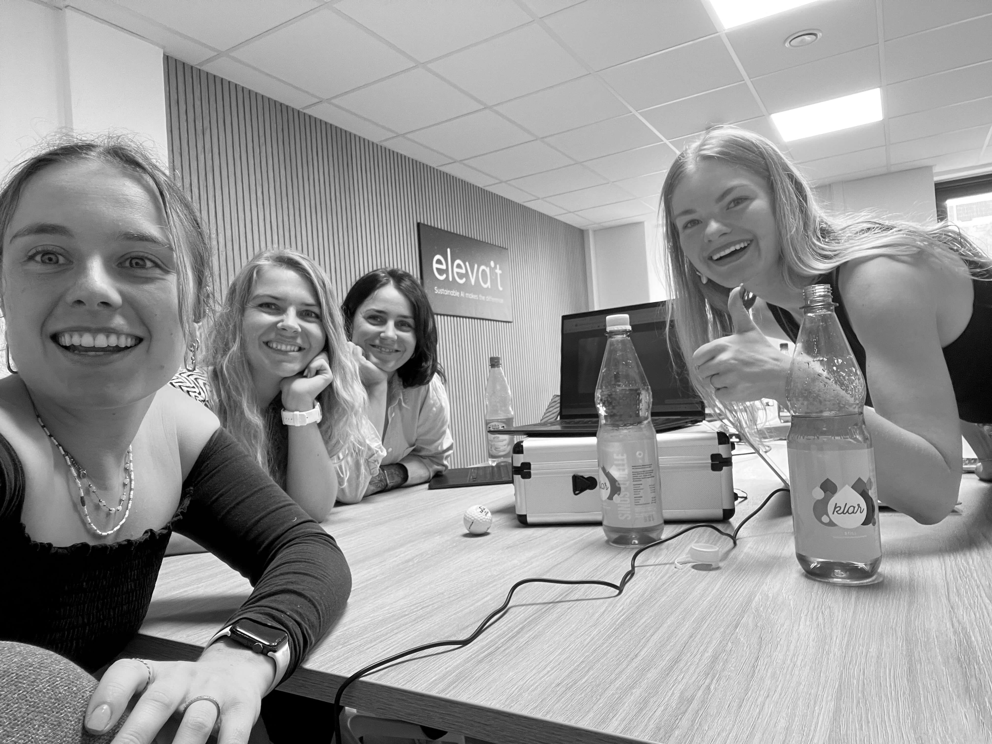 Vier junge Frauen sitzen an einem Tisch vor einem Laptop, lachen und zeigen Daumen hoch in einem modernen Büro.