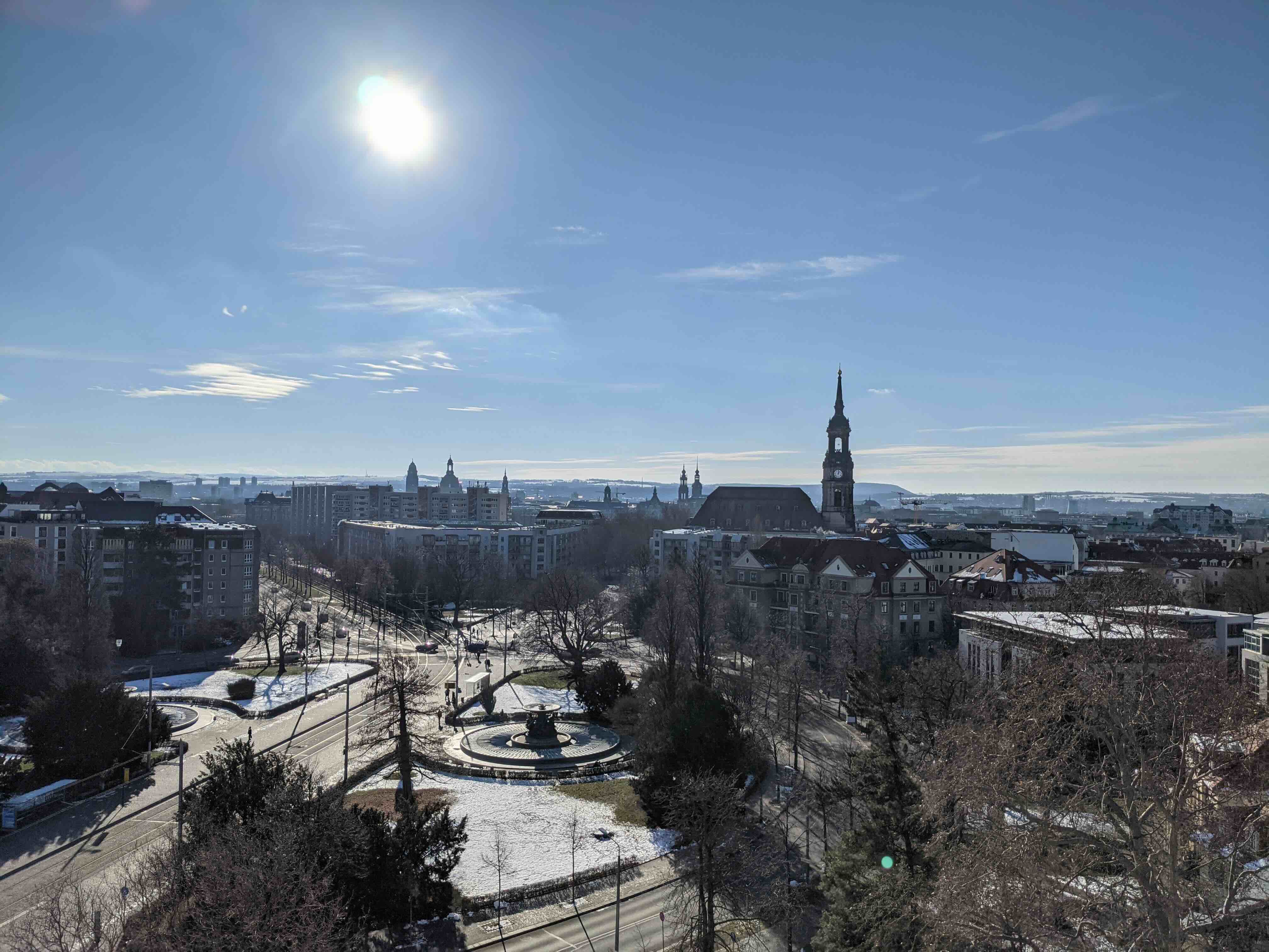 Stadtpanorama bei sonnigem Himmel mit einer großen Kirche, einer runden Verkehrsinsel und verschneiten Flächen.