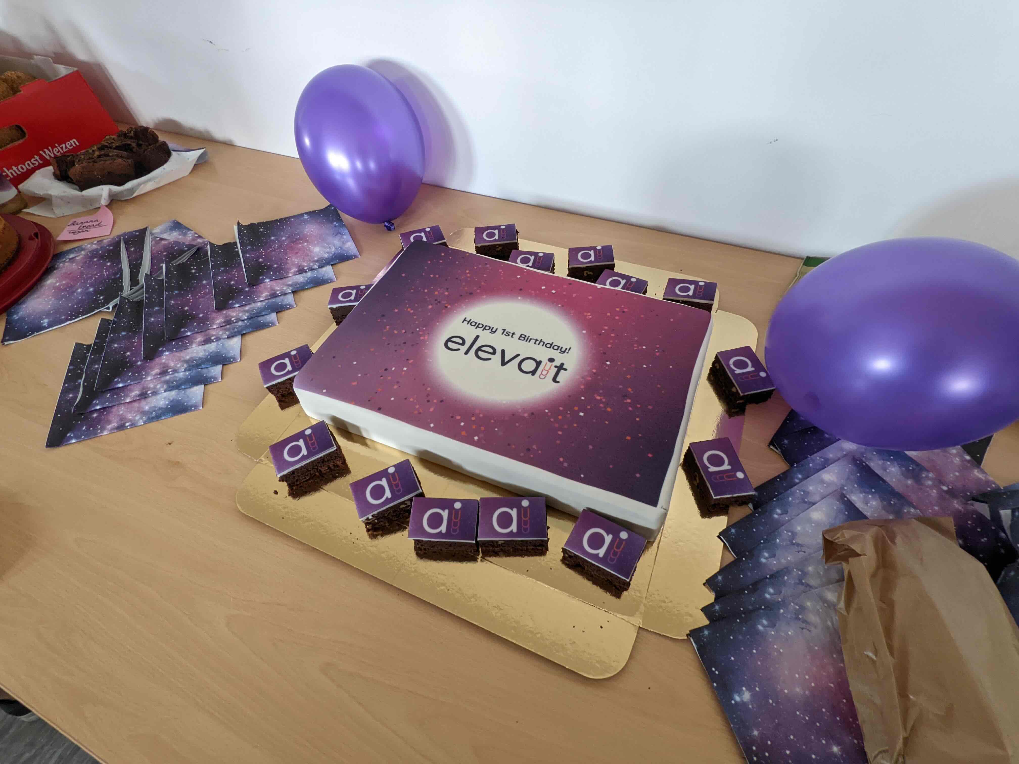 Geburtstagskuchen mit Aufschrift ‚Happy 1st Birthday! elevait‘, umgeben von kleinen Kuchenstücken mit lila Auflegern und lila Luftballons auf einem Holztisch.
