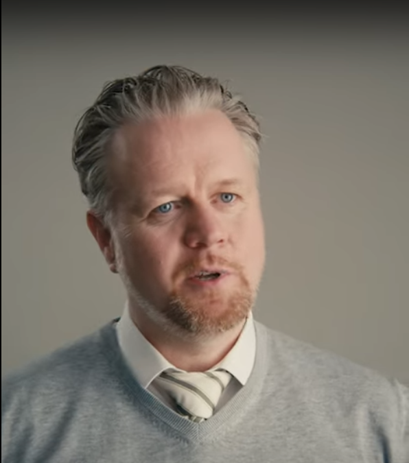 Man with blue eyes, light brown hair, and a trimmed beard wearing a gray sweater over a white shirt and striped tie against a plain gray background.