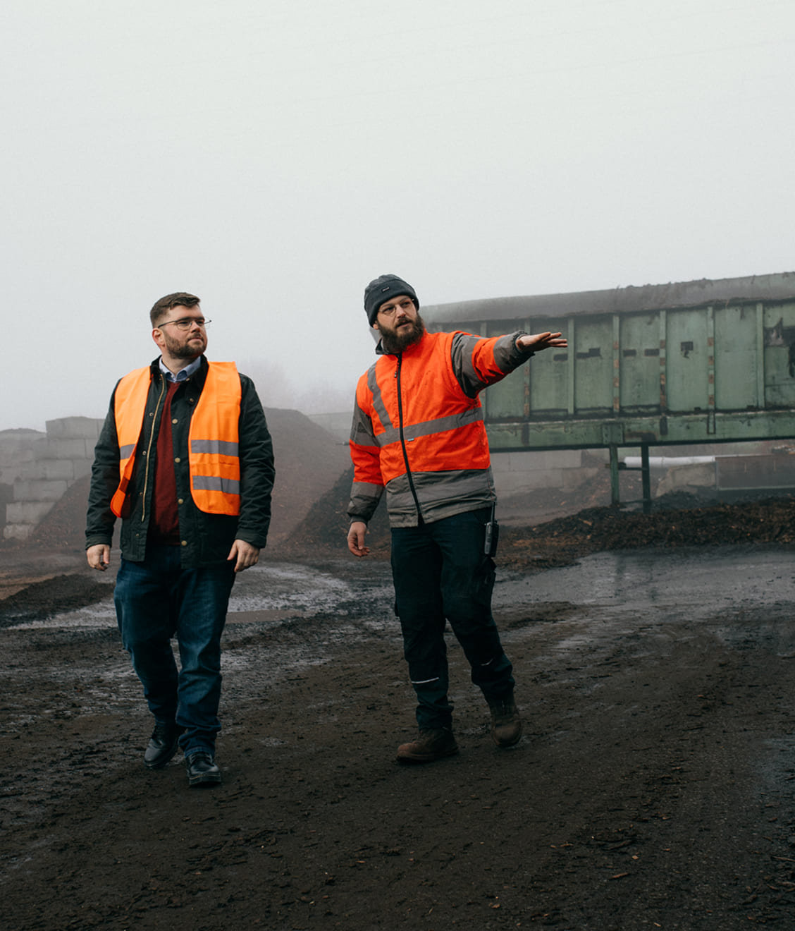 Two men wearing orange safety vests walking on a muddy construction or industrial site, one pointing ahead.