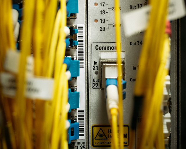 Close-up of yellow fiber optic cables connected to a network patch panel with blue connectors and warning labels.