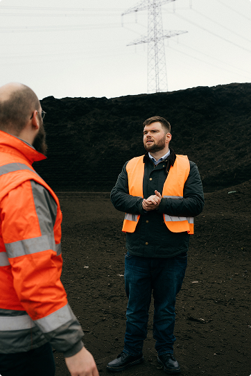 Two men in orange safety vests standing and talking outdoors near a large pile of dark soil or coal with power lines in the background.