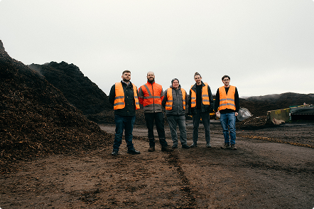 Five men wearing orange safety vests standing on a dirt ground with large piles of dark material in the background under a cloudy sky.