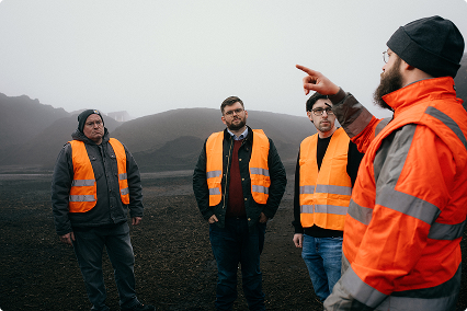 Worker in orange safety jacket instructing three men wearing orange safety vests at a foggy outdoor site.