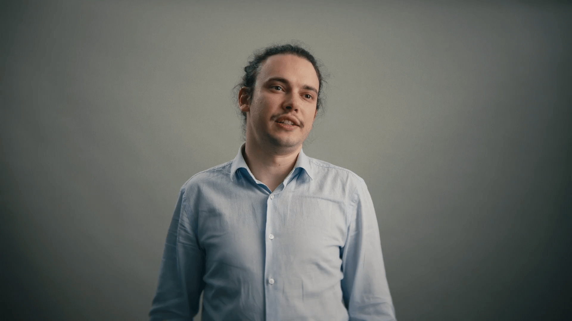 Junger Mann mit zusammengebundenem Haar und hellblauem Hemd vor einem grauen Hintergrund.