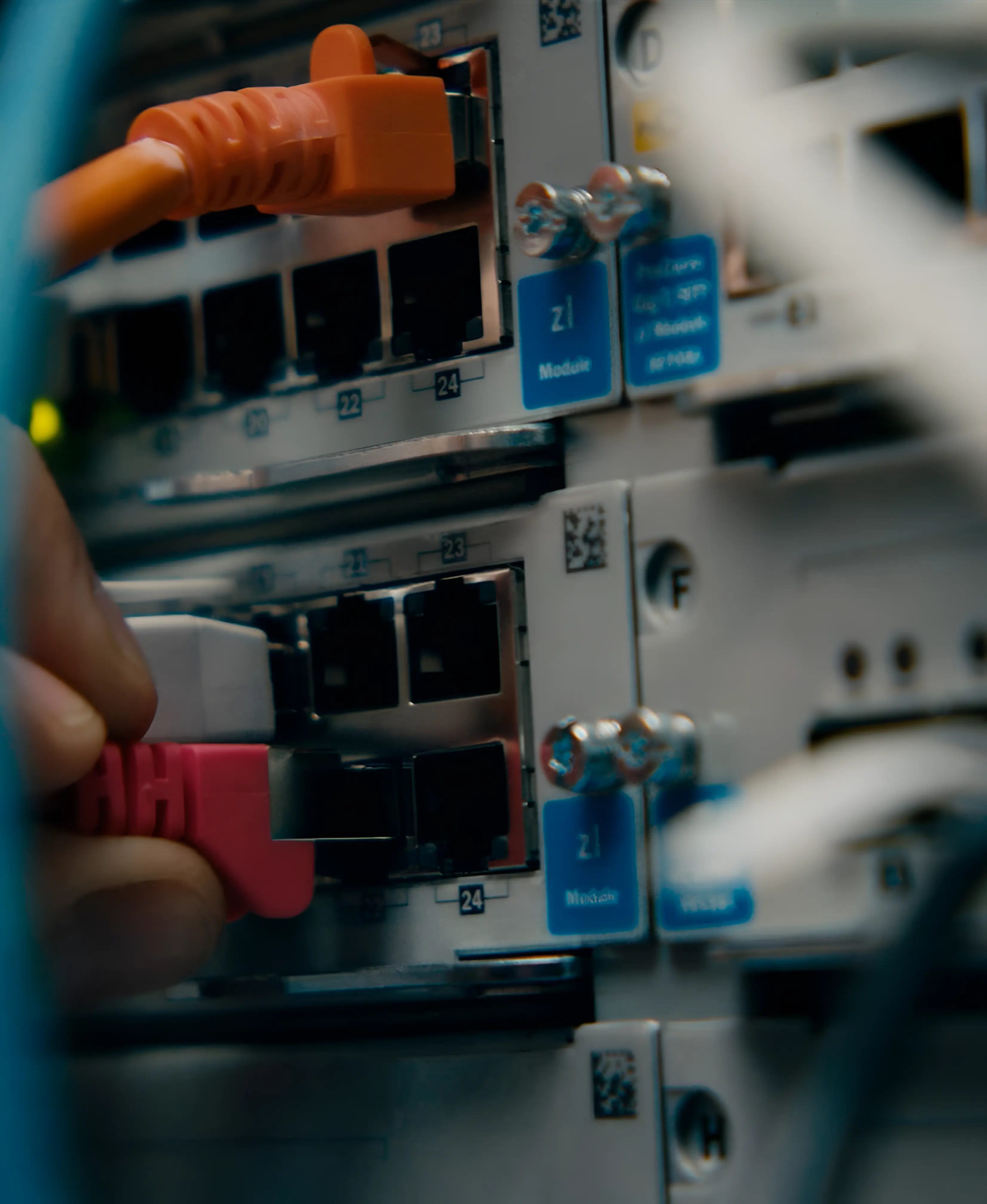 Hand plugs a pink network cable into a port on a network switch with an orange cable connected above.