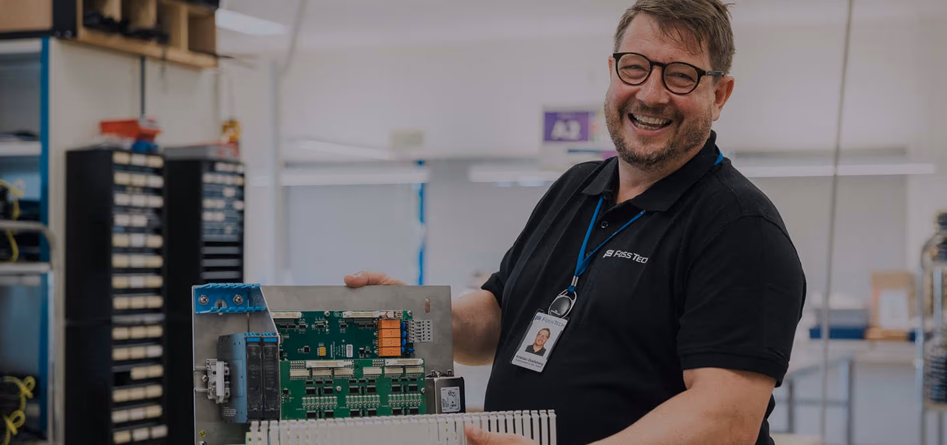 Smilende mann med briller holder et stort kretskort i et verksted.