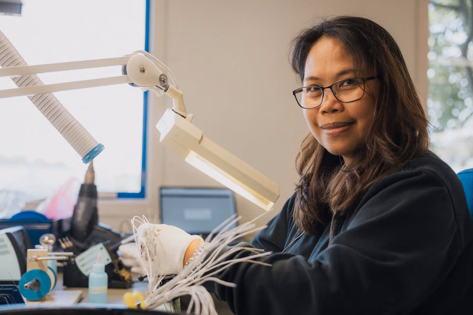 Kvinne med briller som arbeider med tråd og verktøy ved et arbeidsbord med lampe i et lyst rom.
