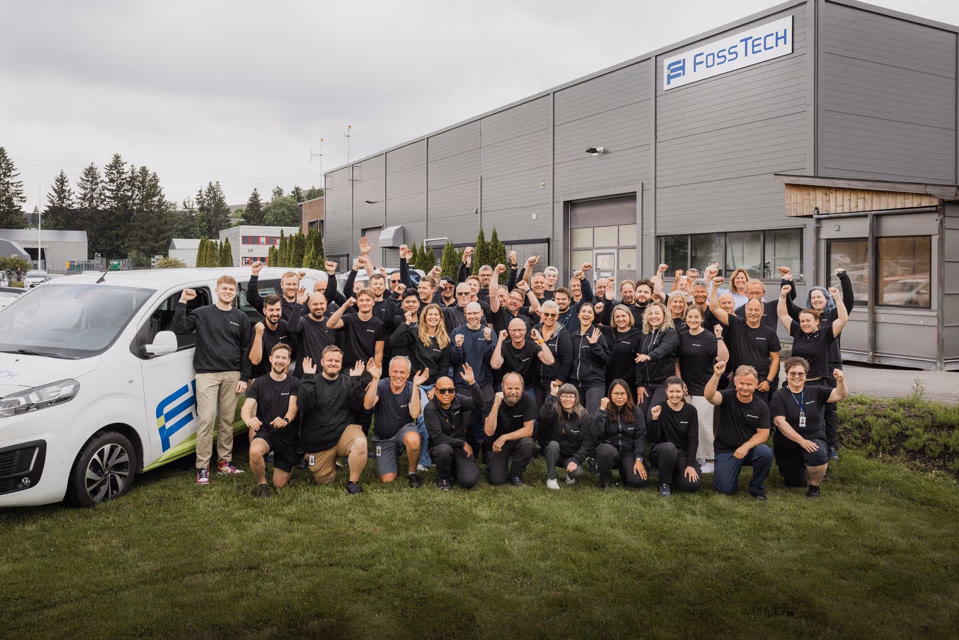 En stor gruppe ansatte i svarte klær hever armene foran et Fosstech-bygg og en hvit firmabil på en gressplen.