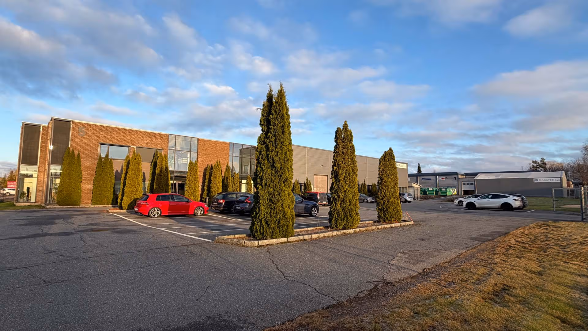 Industrielt bygg med parkeringsplass og flere biler under en blå himmel med spredte skyer.