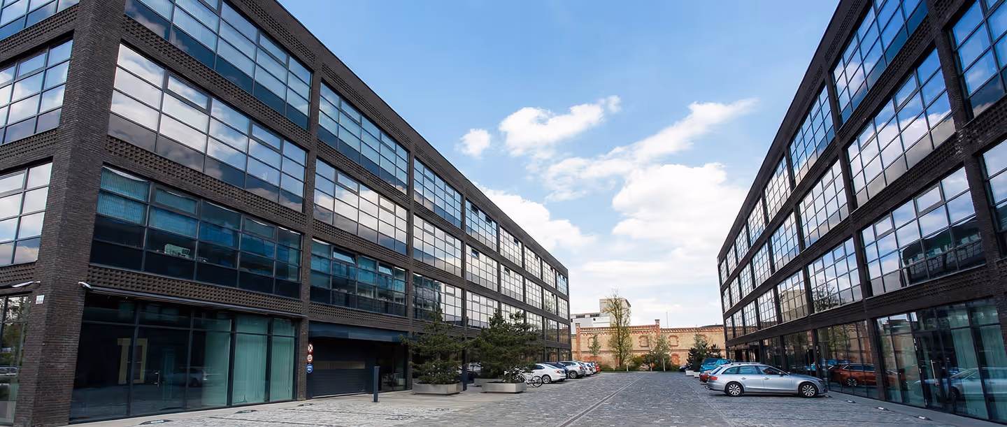Dvouvrstvé kancelářské budovy s velkými skleněnými okny po obou stranách dlážděného parkoviště s několika zaparkovanými auty.