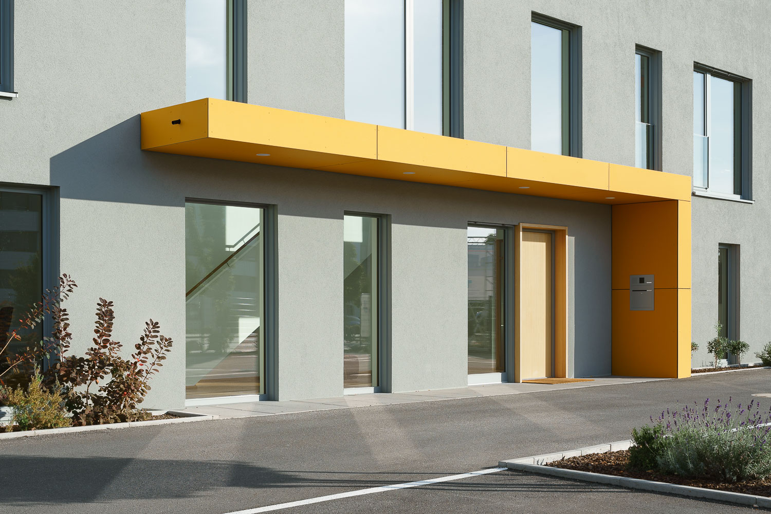 Moderner Gebäudeeingang mit einer gelben Überdachung und großen Fenstern an grauer Fassade.