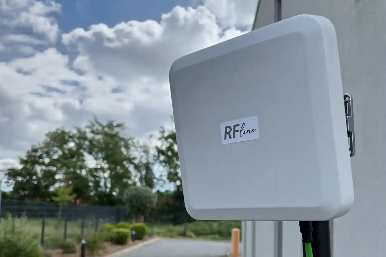 Weiße rechteckige externe RF-Gerät an Hauswand mit bewölktem Himmel und Bäumen im Hintergrund.