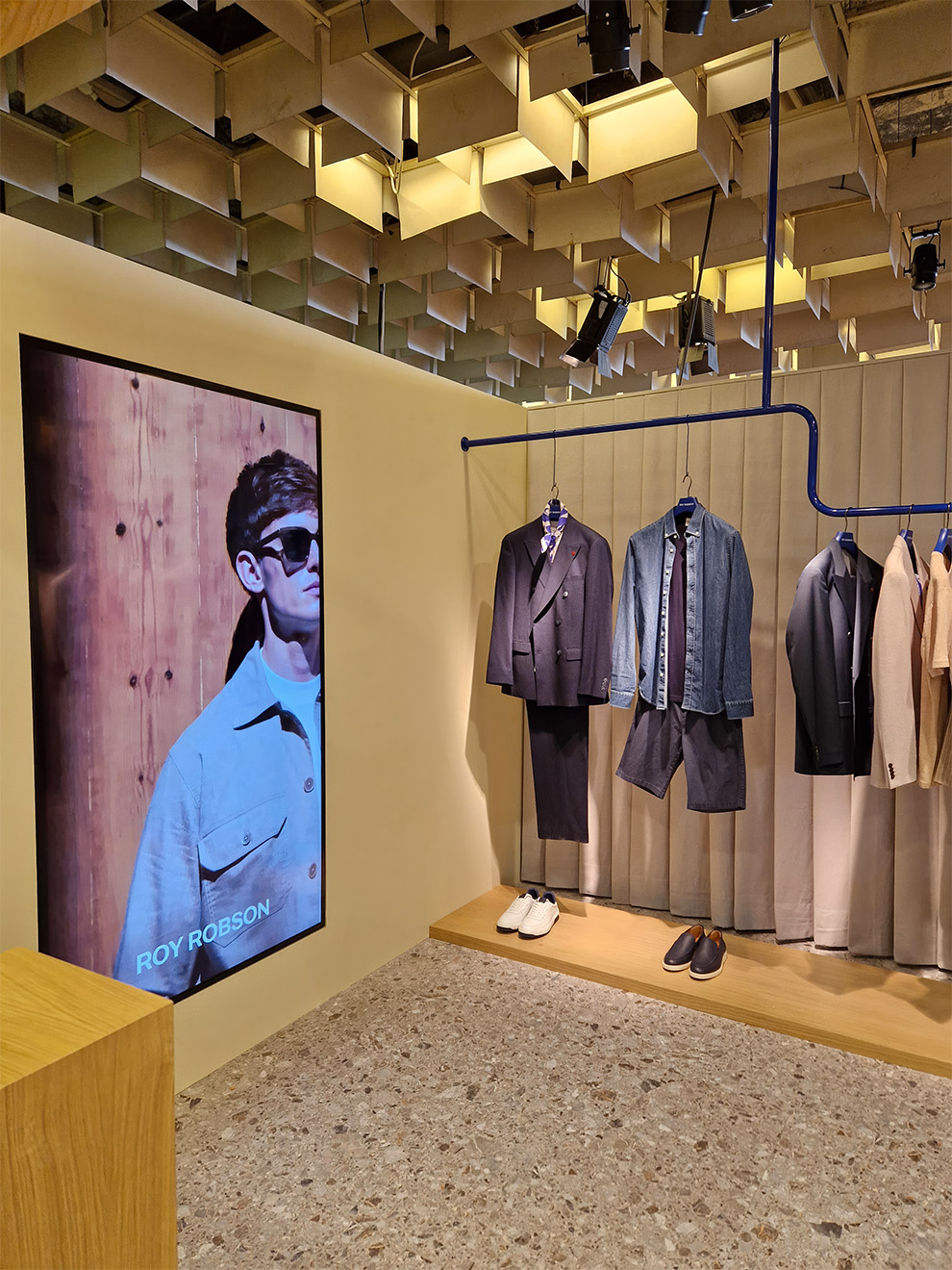 Dettaglio interno dello stand Roy Robson a Pitti Uomo 2024, schermo con visual di campagna e capi appesi su struttura tubolare blu con pedana in legno.