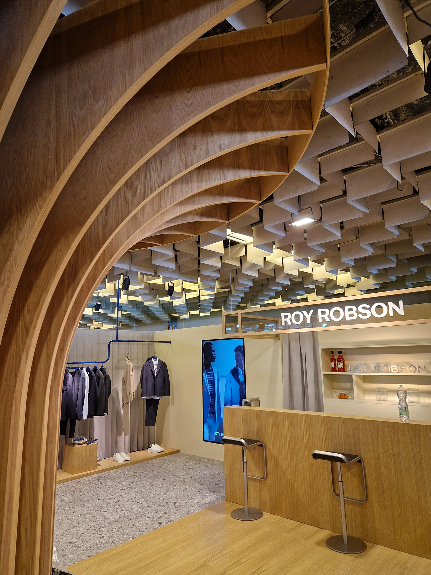 Interno stand Roy Robson a Pitti Uomo 2024, arco in legno a lamelle, bar in rovere con sgabelli e schermo verticale, esposizione capi su struttura tubolare blu.