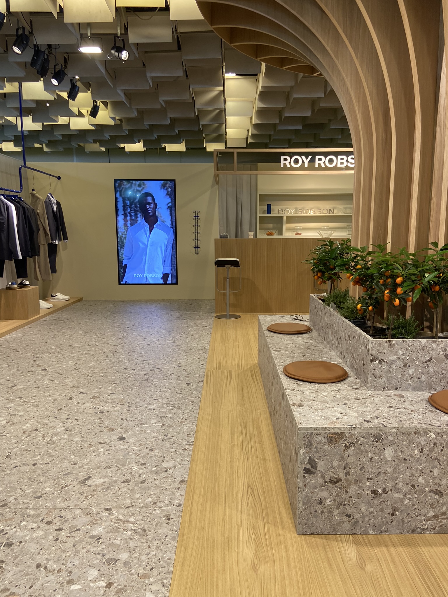 Allestimento Roy Robson a Pitti Uomo 2024, focus sull’isola centrale con agrumi e sedute, pavimento effetto pietra, maxi schermo e bancone hospitality.