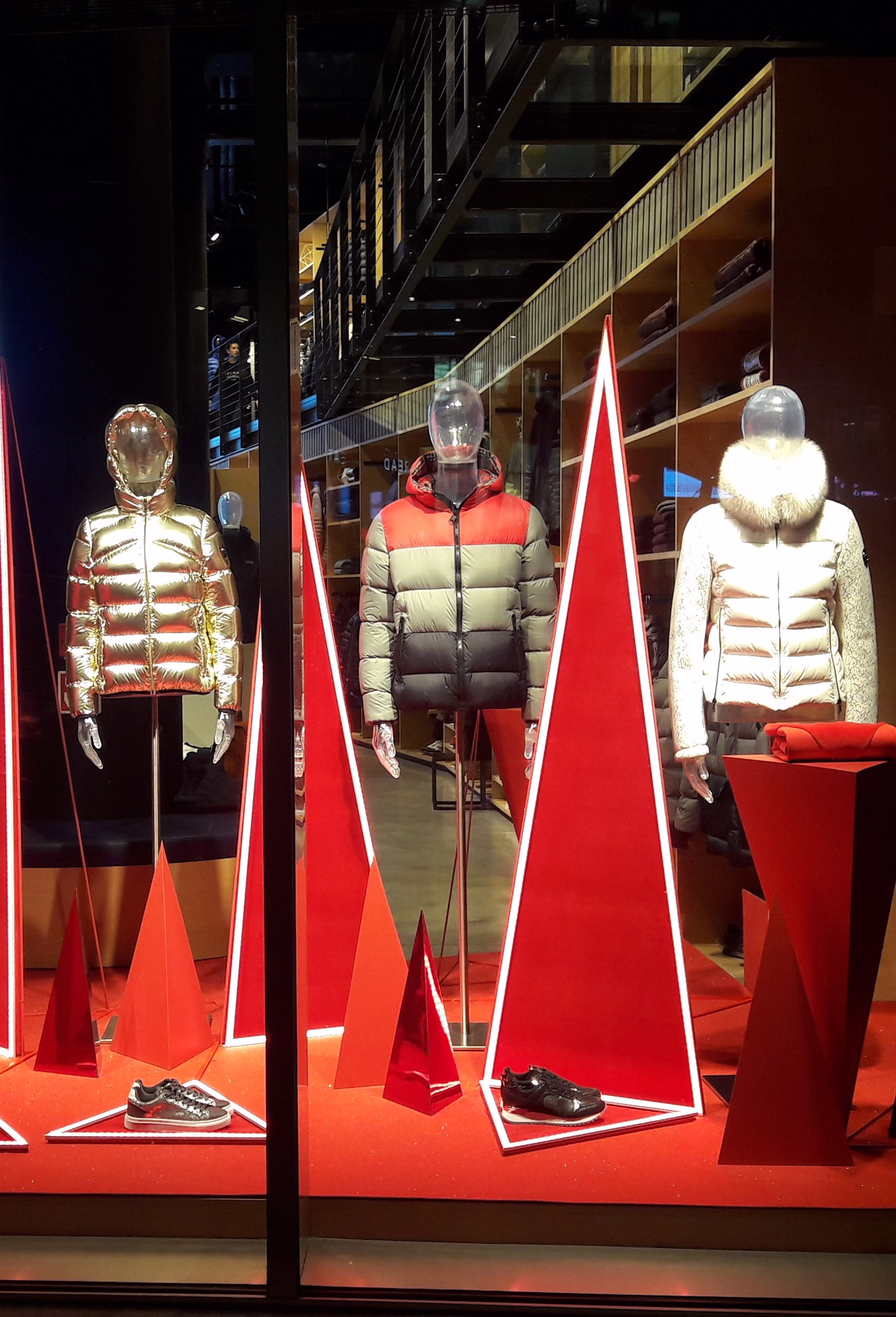 Vetrina Colmar con tre manichini in piumini metallizzati e tricolore, allestimento scenografico con triangoli rossi illuminati e scarpe esposte su pedane.