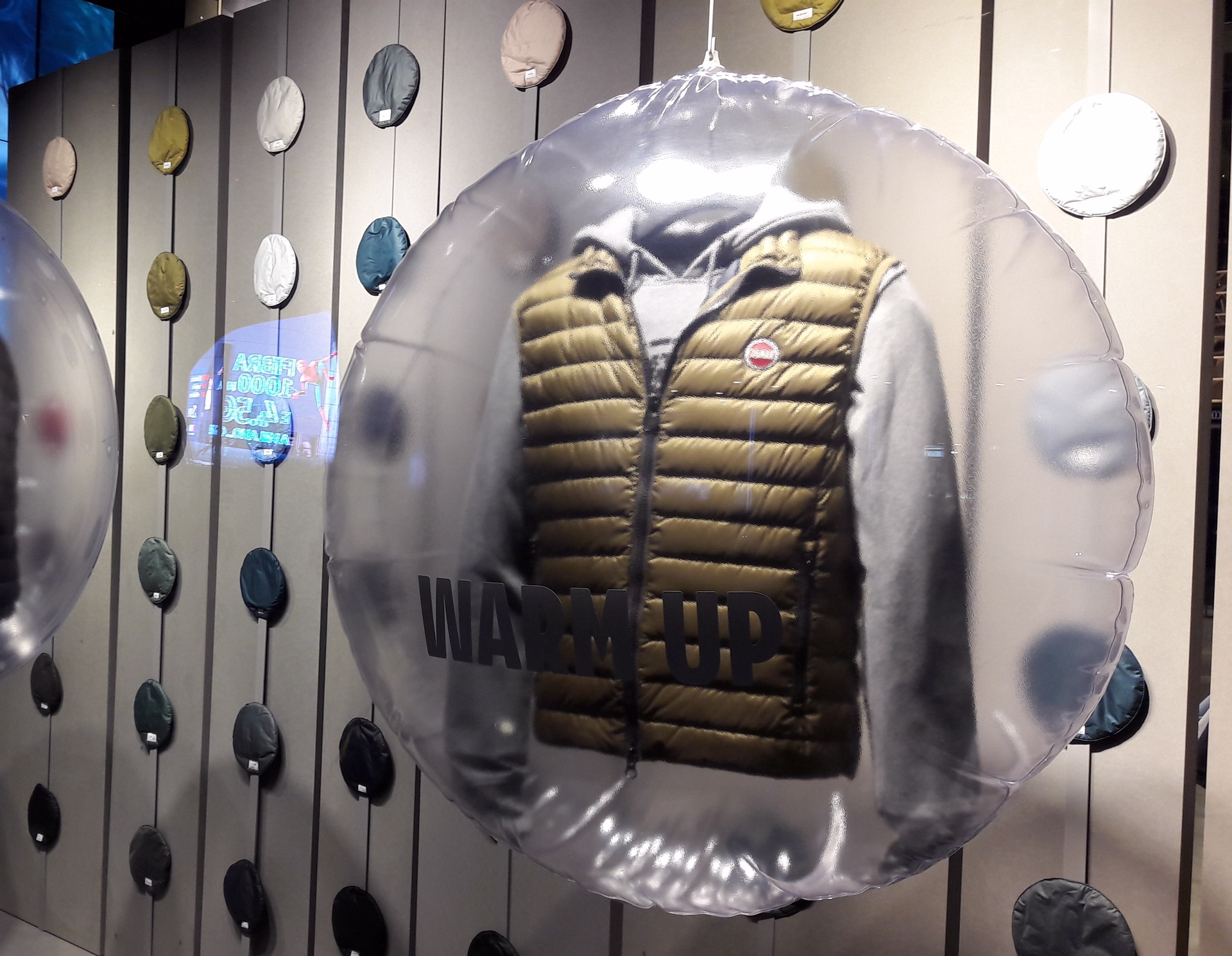 Vetrina con capo imbottito esposto dentro una grande sfera trasparente con scritta “Warm Up”, sullo sfondo parete con dischi colorati appesi.