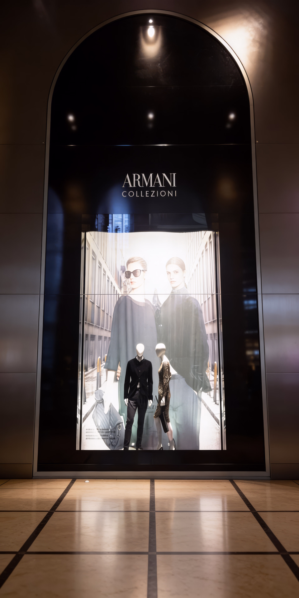 Vetrine Armani con cornici ad arco e loghi di linea, maxi visual di campagna e manichini in outfit esposti su pedane illuminate.