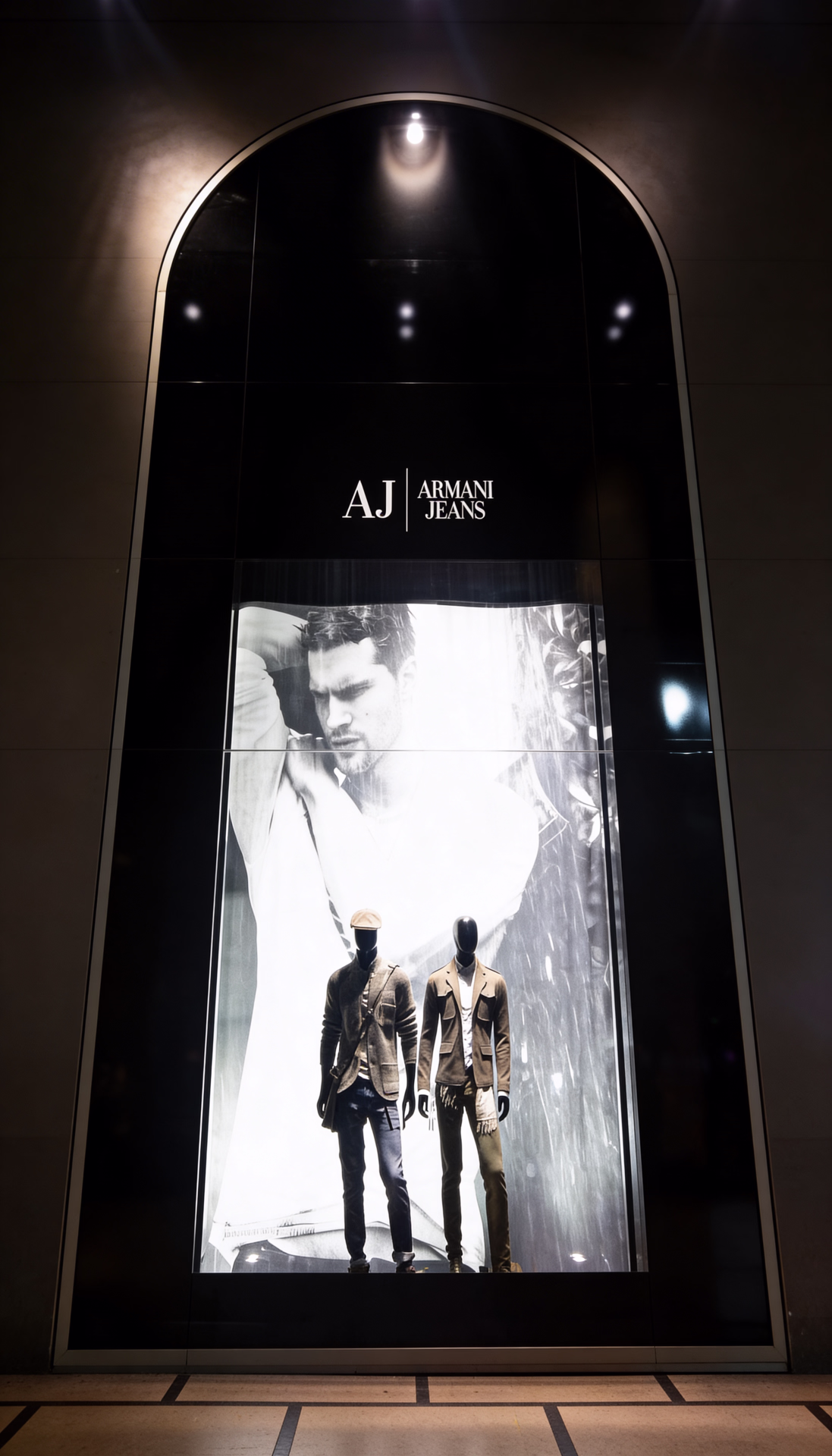 Vetrine Armani con cornici ad arco e loghi di linea, maxi visual di campagna e manichini in outfit esposti su pedane illuminate.