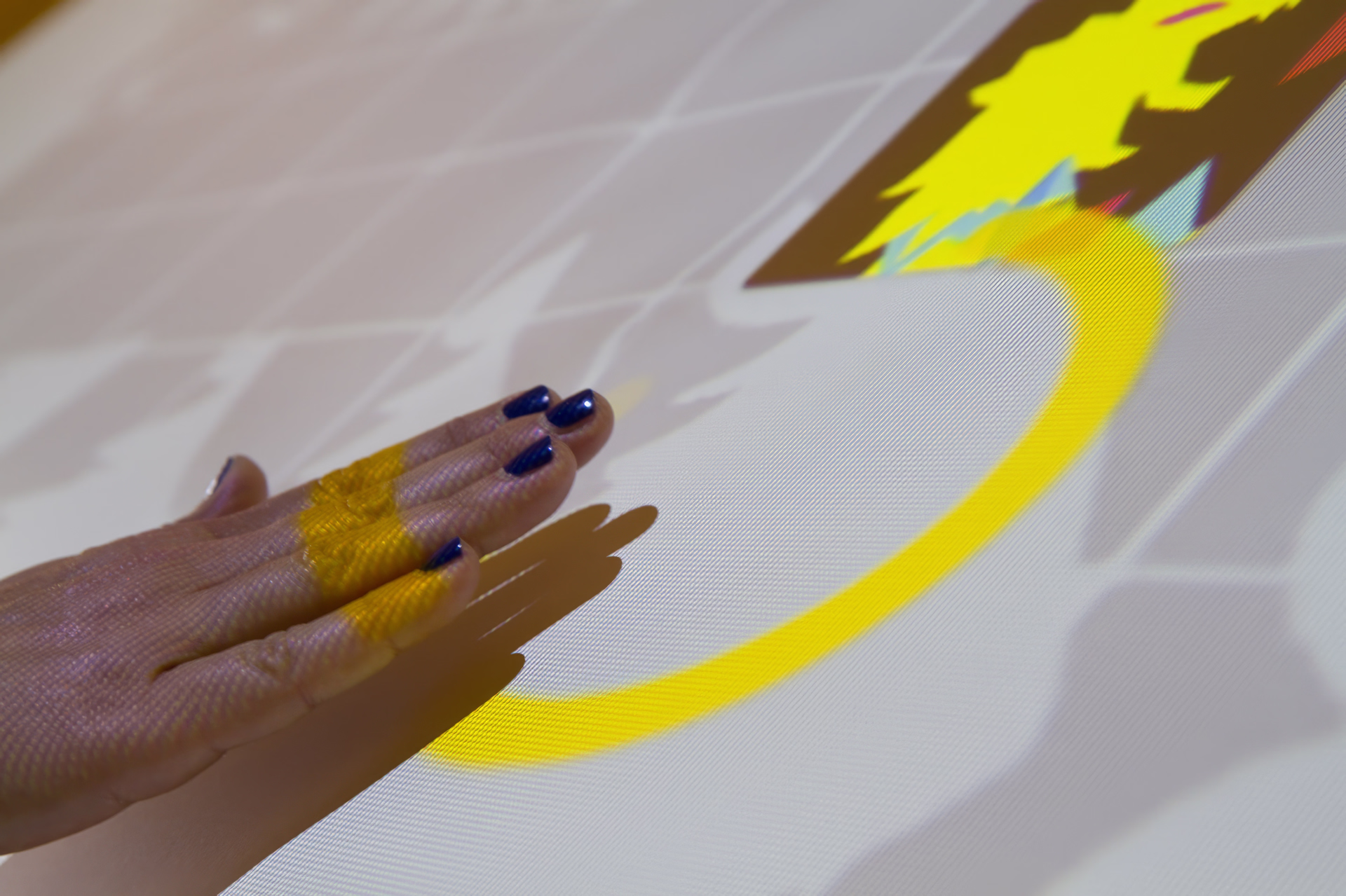 Mano che sfiora una superficie interattiva con grafica proiettata in giallo e bianco, come in un’installazione digitale.
