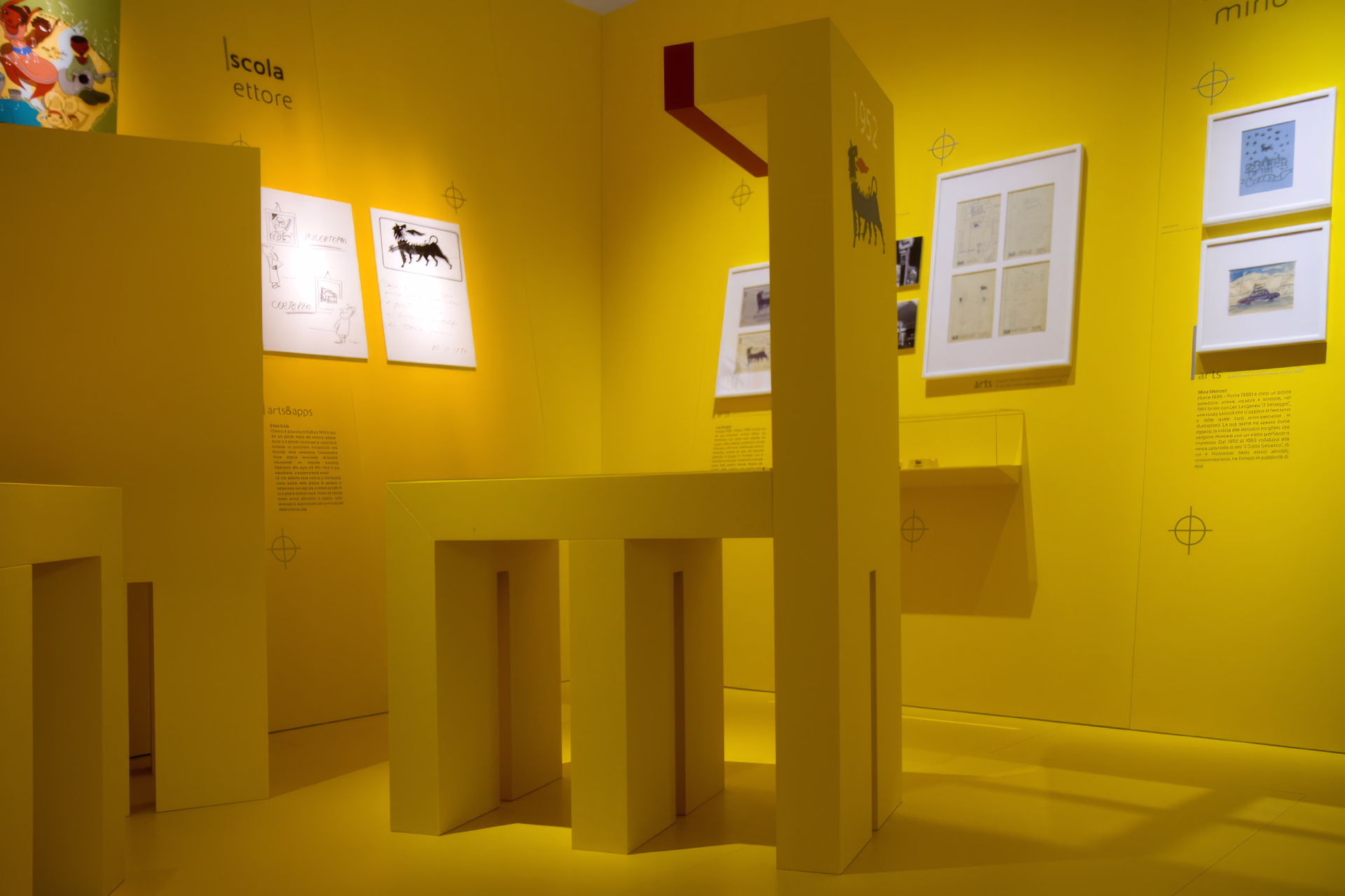 Installazione monolitica gialla con moduli geometrici e dettagli rossi, circondata da disegni, fotografie e pannelli didascalici che raccontano il percorso storico e visivo del brand Eni.