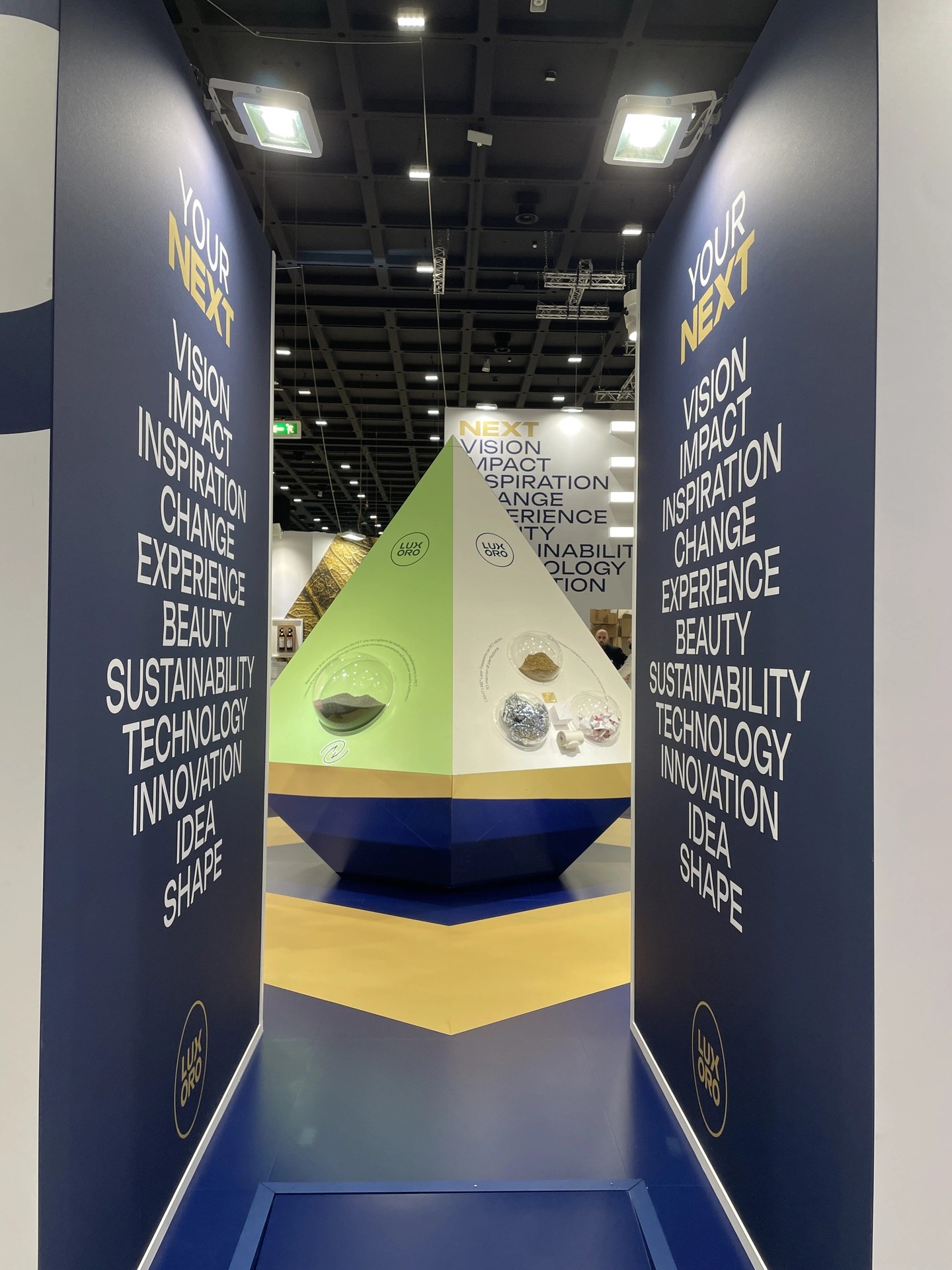 Corridoio di stand fieristico con pareti blu e scritte “YOUR NEXT” e lista di parole, al centro struttura piramidale verde e bianca con logo Luxoro e piccole teche circolari con campioni di materiali.