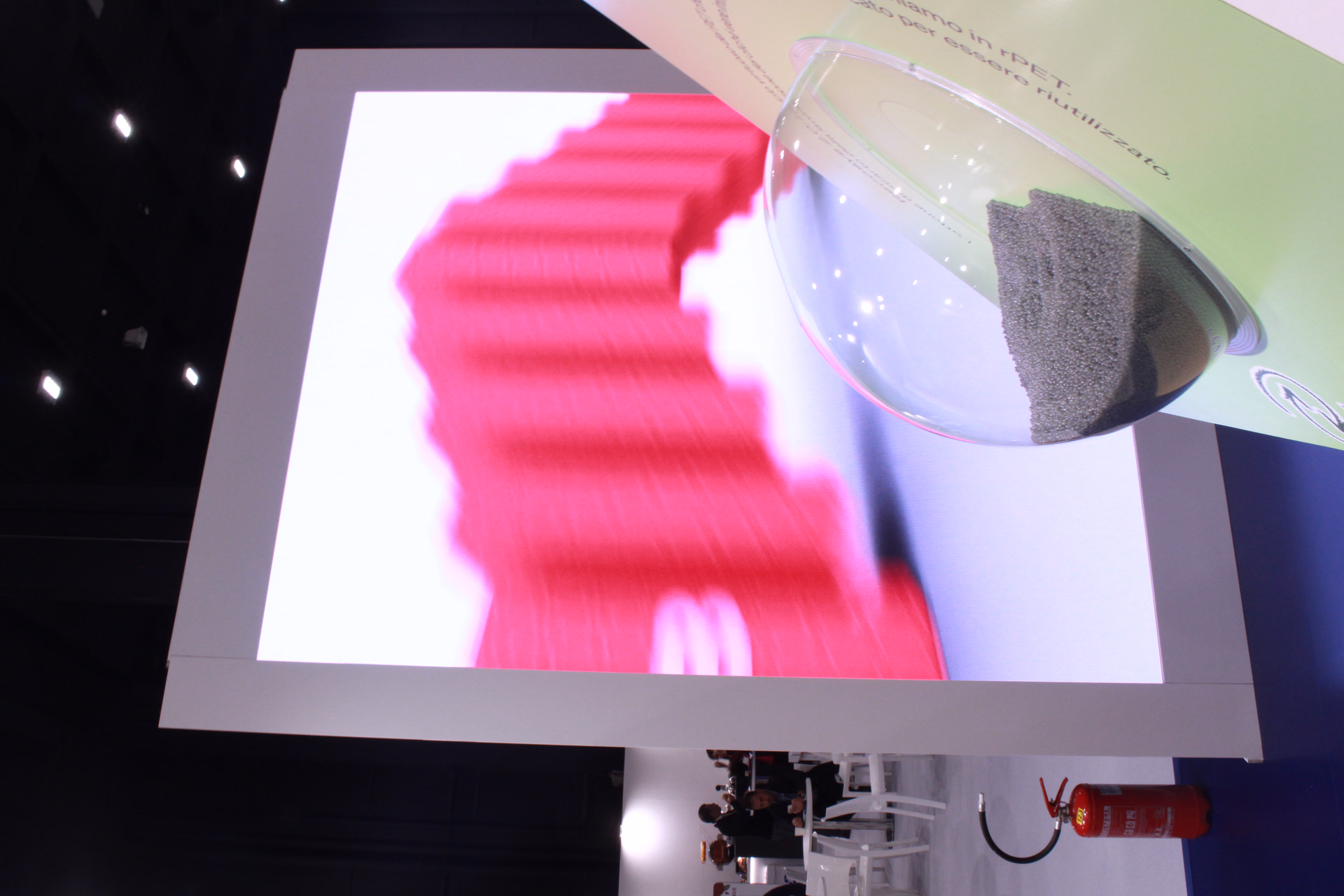 Maxischermo verticale in fiera con video astratto rosso, in primo piano una teca trasparente a cupola con granuli scuri su pannello inclinato, sullo sfondo area lounge con sedute.