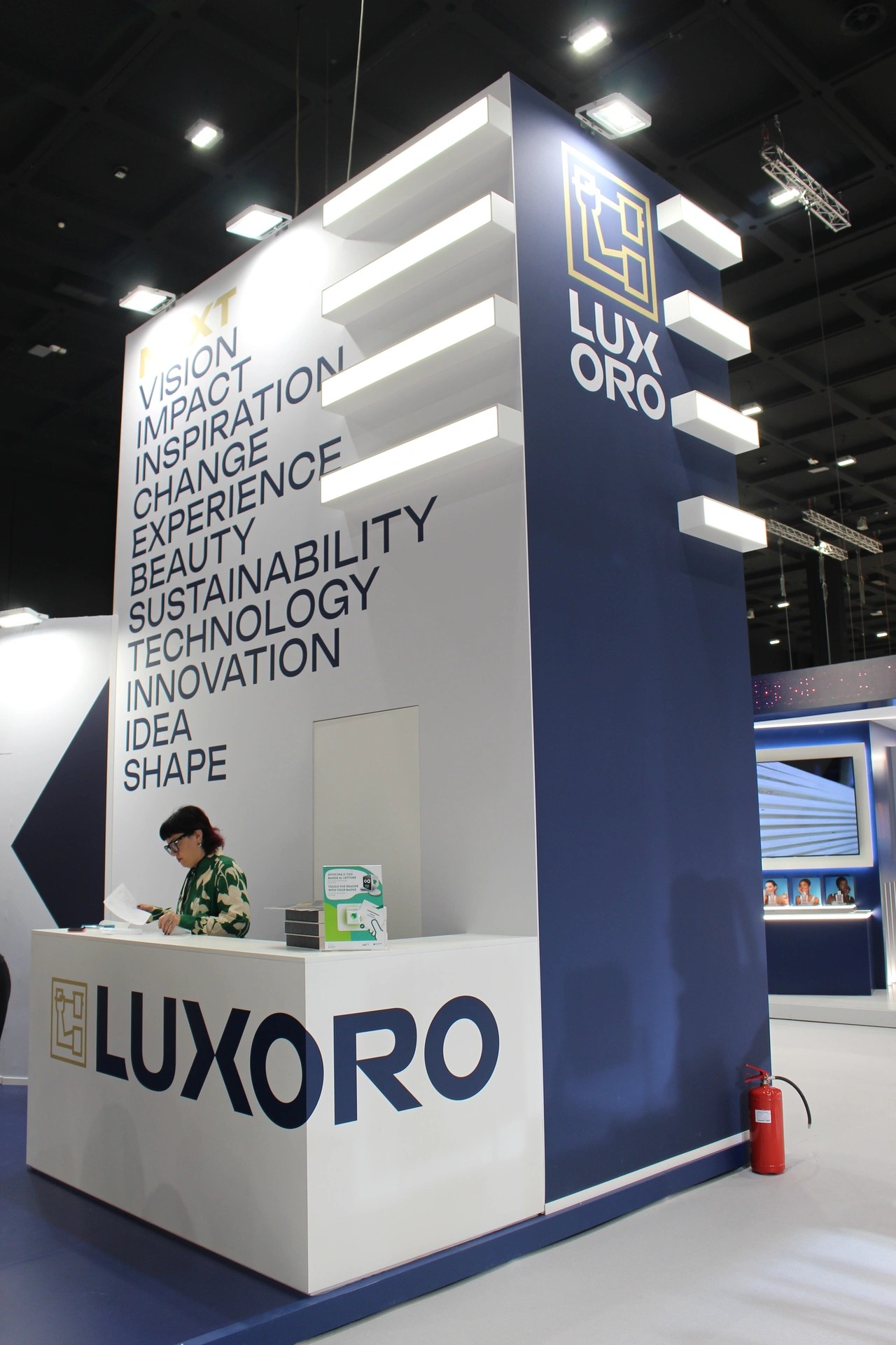 Torre scenografica bianco e blu con logo Luxoro e mensola luminose a sbalzo, parete con parole chiave del brand, desk reception frontale con maxi logotipo e operatore in accoglienza.