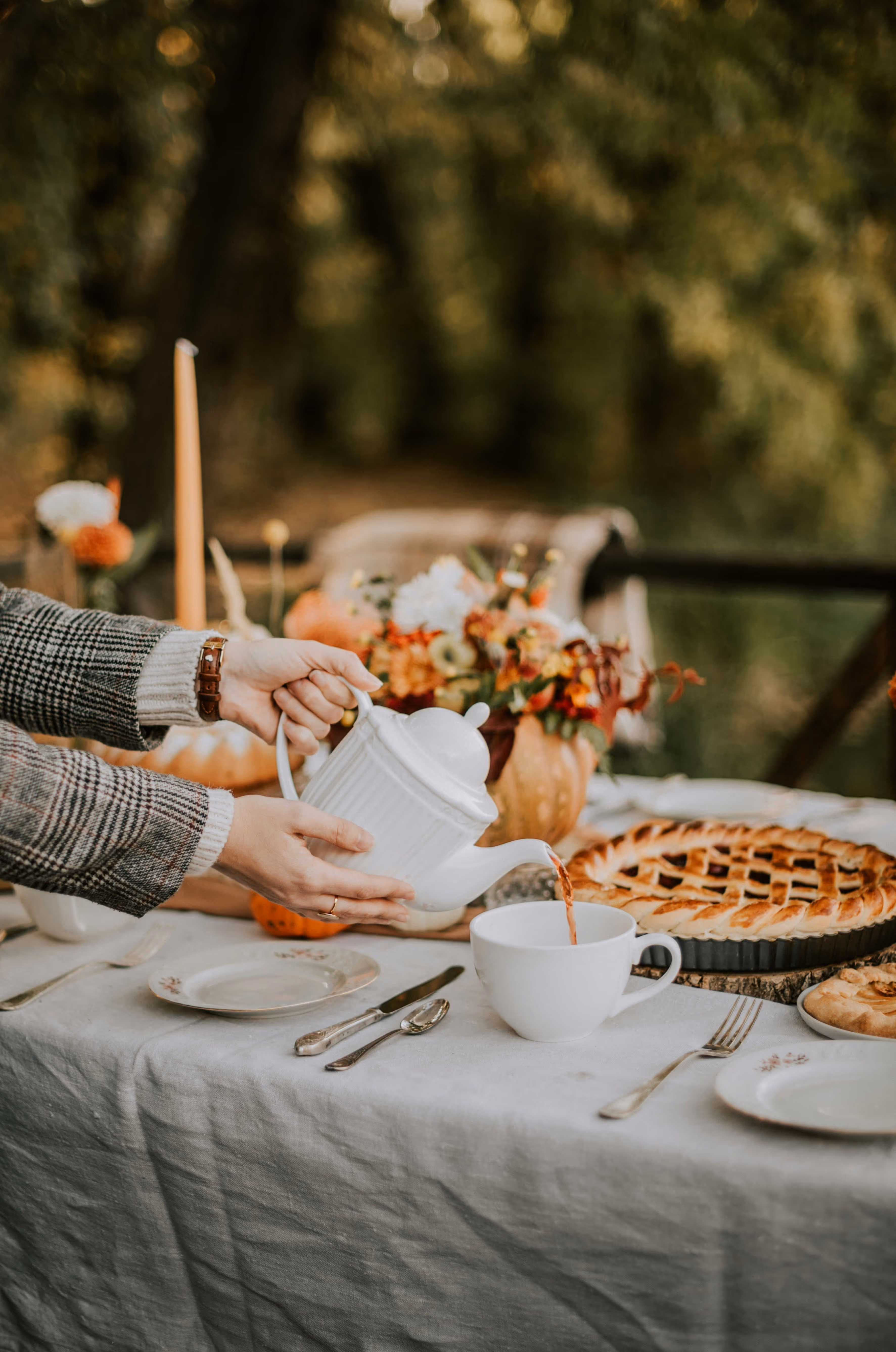 Person pouring tea from a white teapot into a large white cup on a table set with pies, plates, cutlery, and autumn floral decorations.