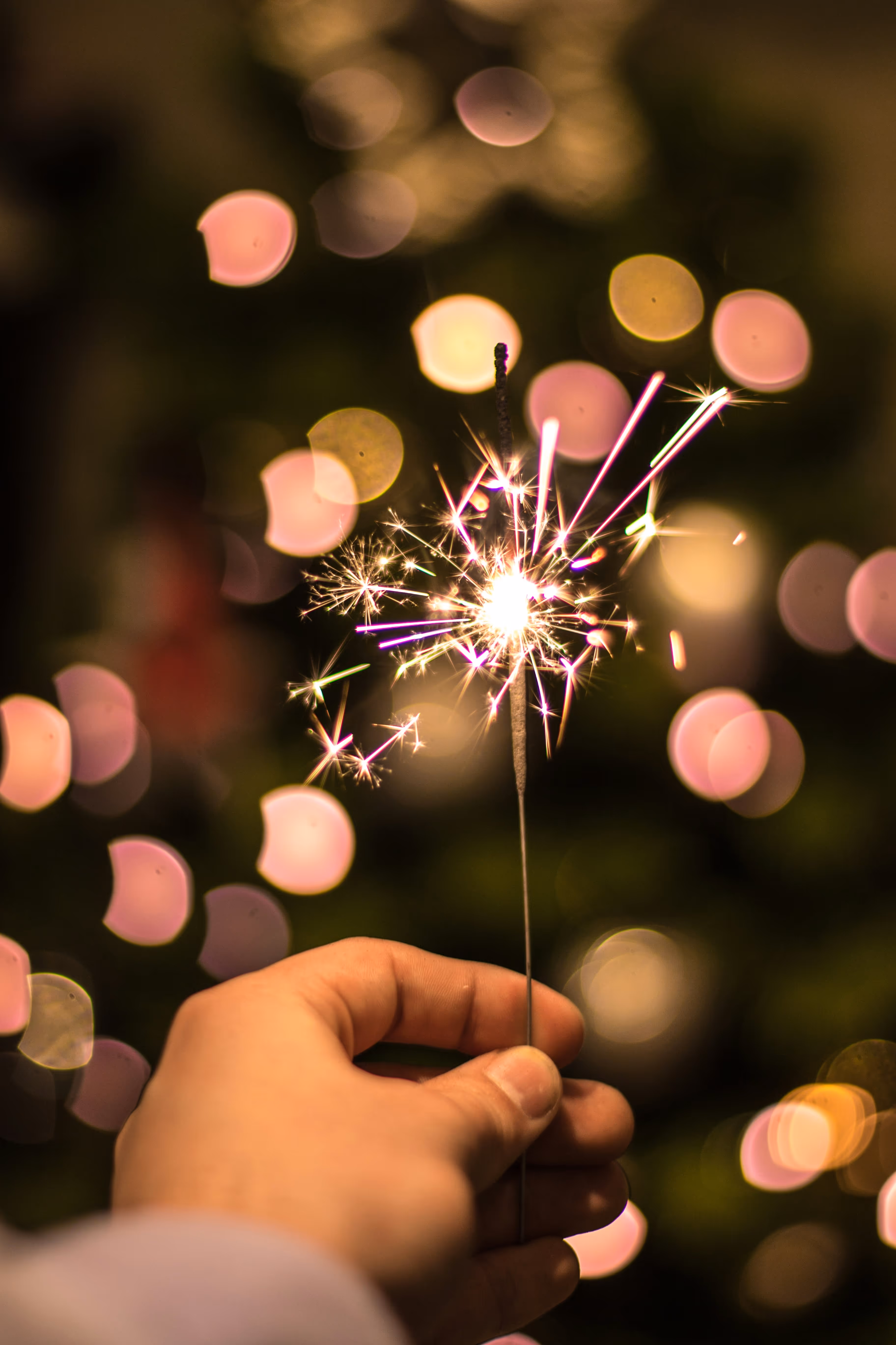 Hand holding a lit sparkler with blurred pink and yellow festive lights in the background.