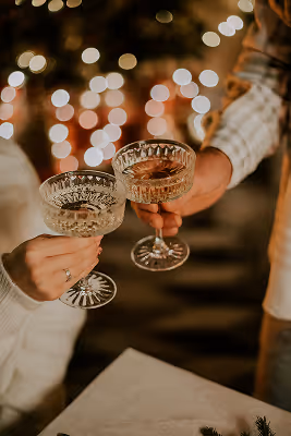 Two people clinking crystal champagne glasses with warm bokeh lights in the background.