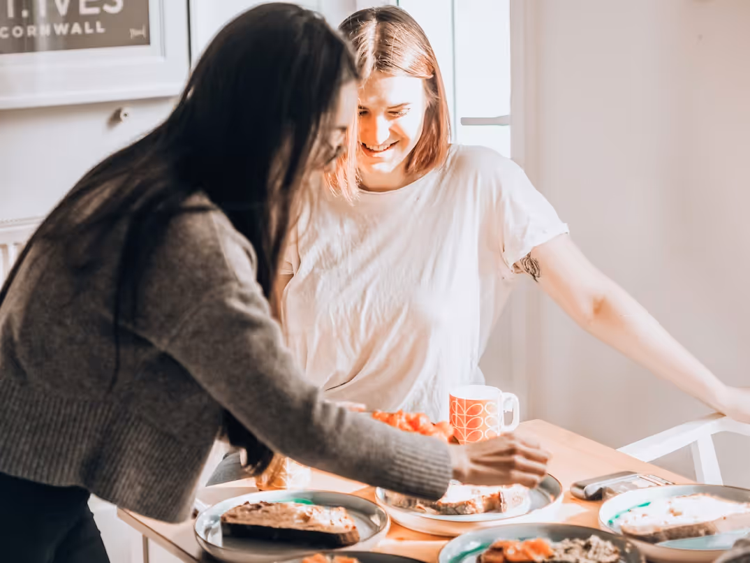 Two women preparing and serving food at a dining table with plates of bread and a beverage mug.