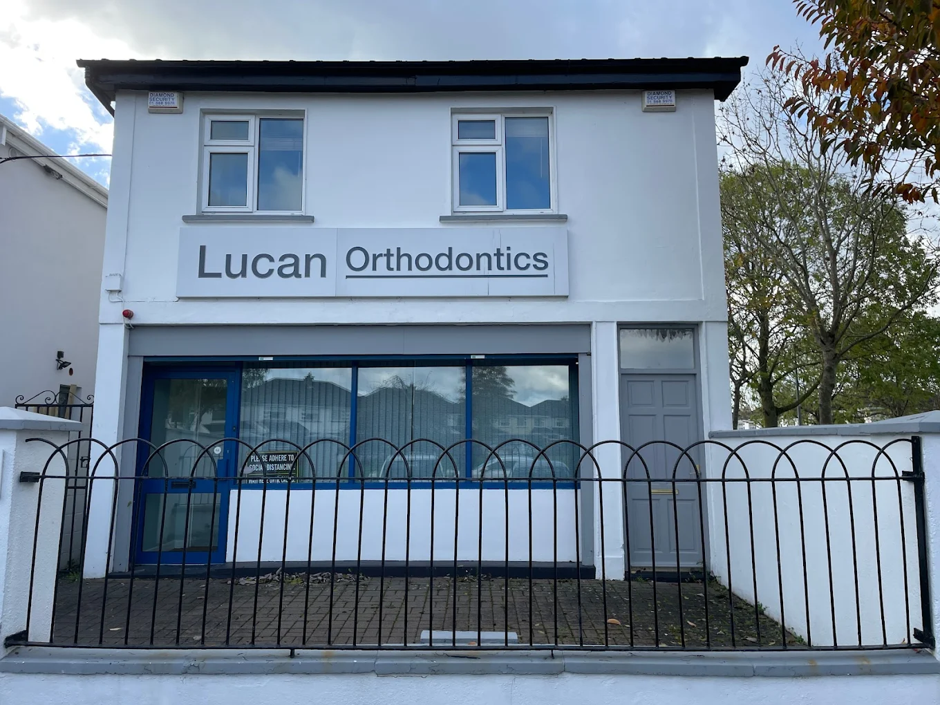 Two-story white building with a sign reading 'Lucan Orthodontics' above a blue door and window, enclosed by a black metal fence.