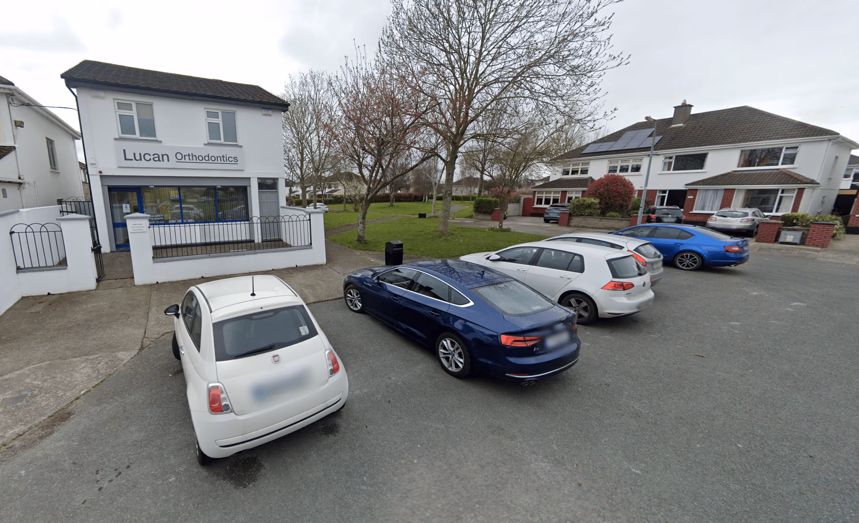 Small parking lot with five cars in front of a white building labeled Lucan Orthodontics and residential houses in the background.