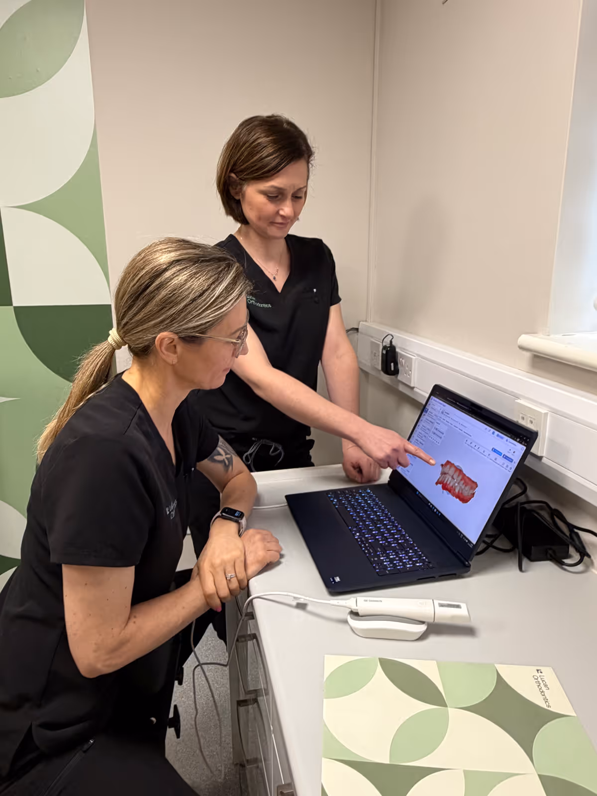 An orthodontist in purple scrubs is showing a dental X-ray on a monitor to a patient seated in a dental chair.