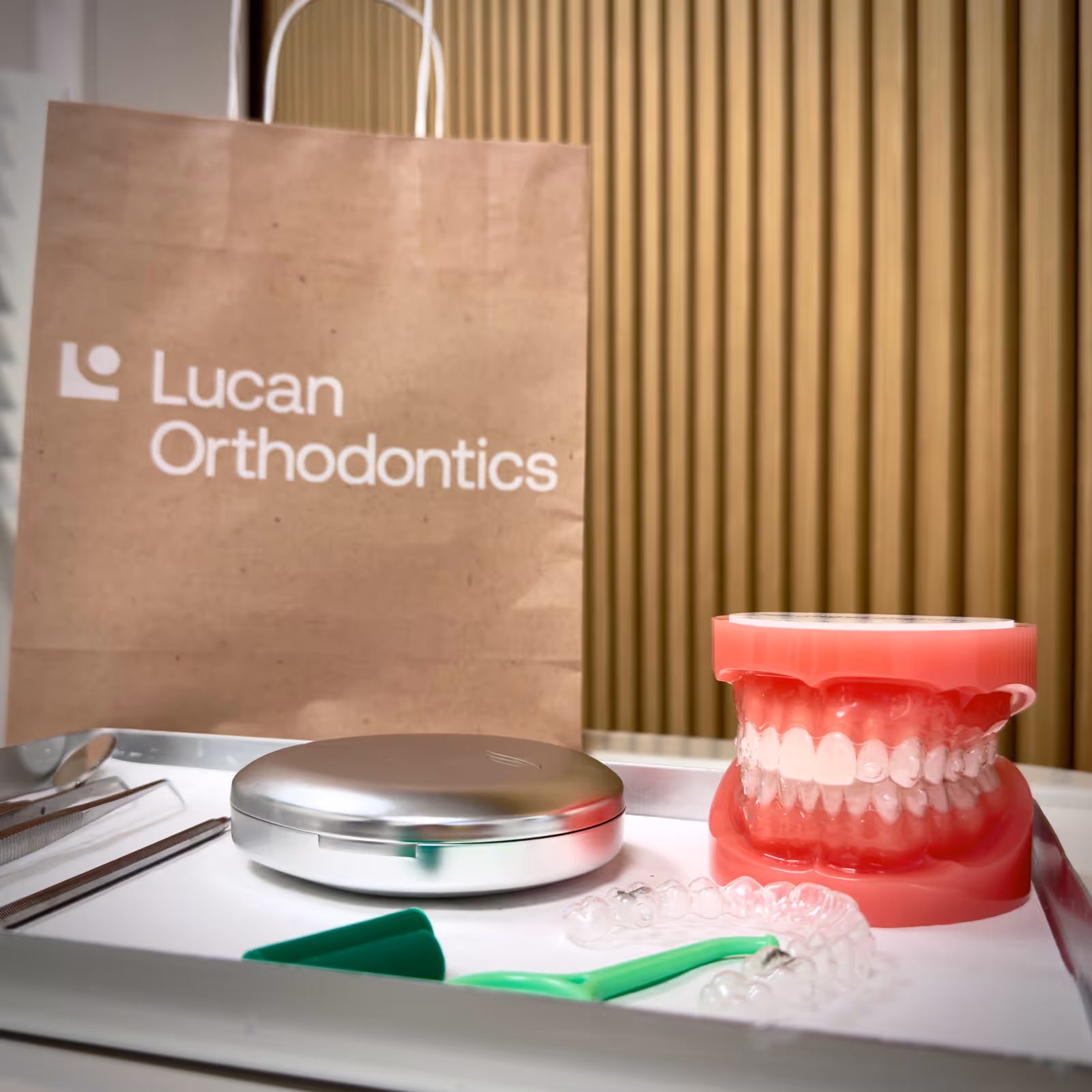 Orthodontic tools including a dental model with clear aligners, a metal case, and dental instruments arranged on a tray with a Lucan Orthodontics branded paper bag in the background.
