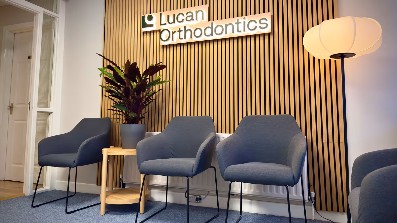Waiting area with three grey chairs, a small round wooden table with a potted plant, a striped wooden wall panel with illuminated Lucan Orthodontics sign, and a tall floor lamp.