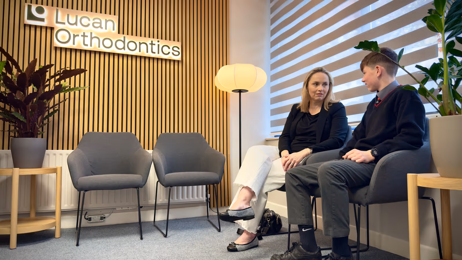 A woman and a teenage boy sitting and talking in a modern waiting room with gray chairs, plants, and a sign that says Lucan Orthodontics.