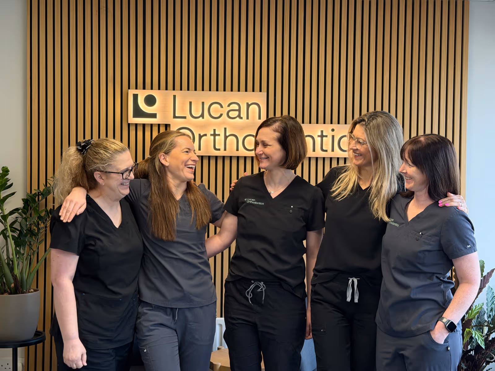 Five women wearing black and gray scrubs standing arm in arm smiling in front of a wooden slat wall with a Lucan Orthodontics sign.